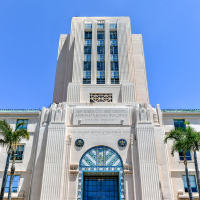 A tall, Art Deco-style building with a blue sky backdrop, featuring palm trees and decorative architectural elements.